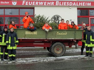 Traktoranhänger mit Weihnachtsbäumen beladen vor dem Feuerwehrgerätehaus, auf dem Anhänger sitzen neben den Tannenbäumen die Jugendfeuerwehrmitglieder, neben dem Anhänger stehen vier aktive Feuerwehrkameraden